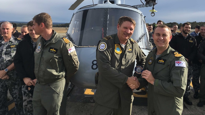 A group of Navy pilots in khaki flight suits standing in front of a grey helicopter.