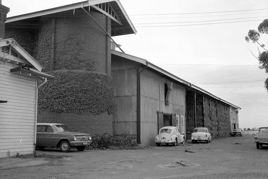 A black and white photo of sheds, with vintage cars parked in front. 