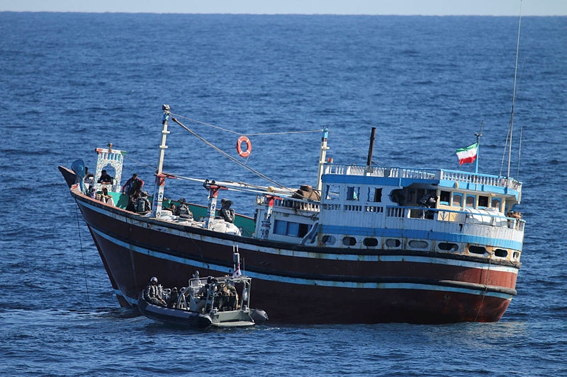 A small military boat floats next to a larger, rustic-looking fishing boat in the ocean.