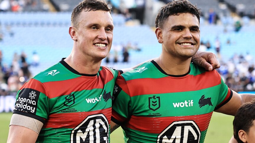 Two men in red and green striped guernseys smile at a camera while standing in a stadium