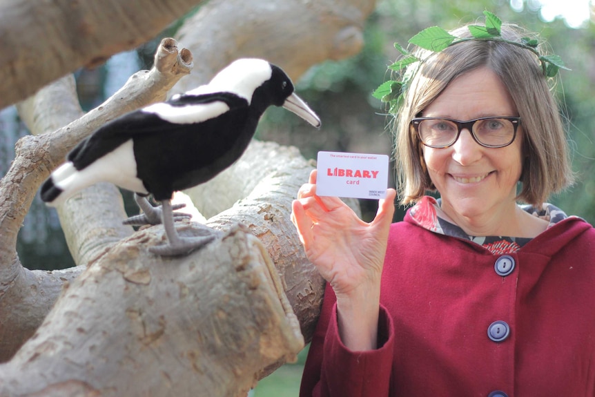 Children's author Ursula Dubosarsky holding up a library card with a stuffed magpie toy to her right.