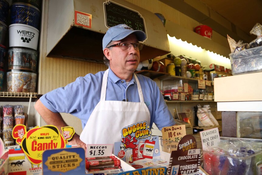 A man in apron and baseball cap stands behind the counter of a lolly store.
