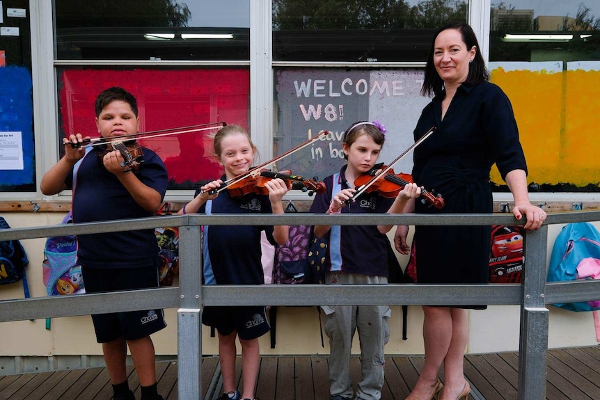 A woman stands outside a classroom beside three primary-aged students, in school uniform, playing the violin.
