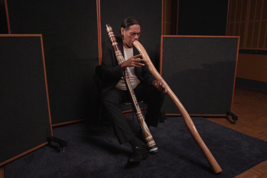 Didgeridoo virtuoso William Barton performs yidaki while seated. He has a second yidaki resting across his shoulder.