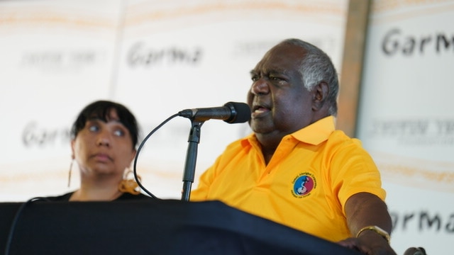 An older Aboriginal man in a yellow shirt speaks into a microphone.