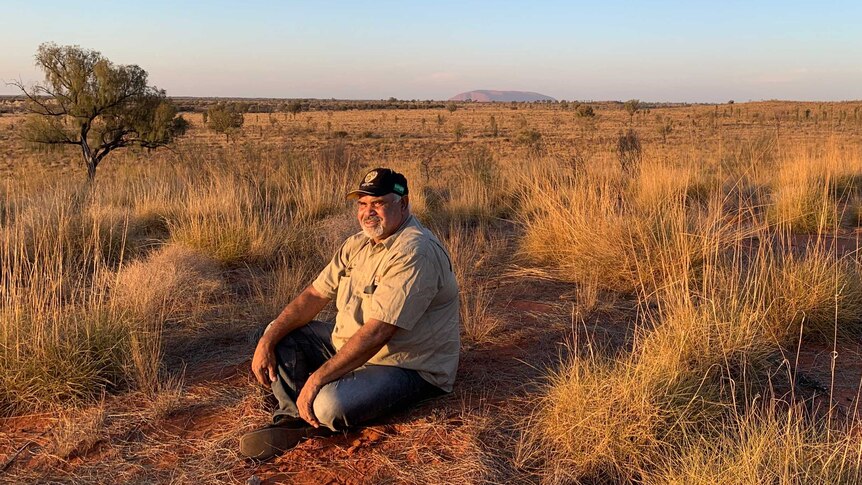 Traditional owner Sammy Wilson sits on red dirt in the sunset with Uluru in the distant background