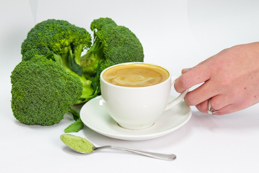 Three bunches of broccoli with a hand holding a cup of coffee and a teaspoon of broccoli powder