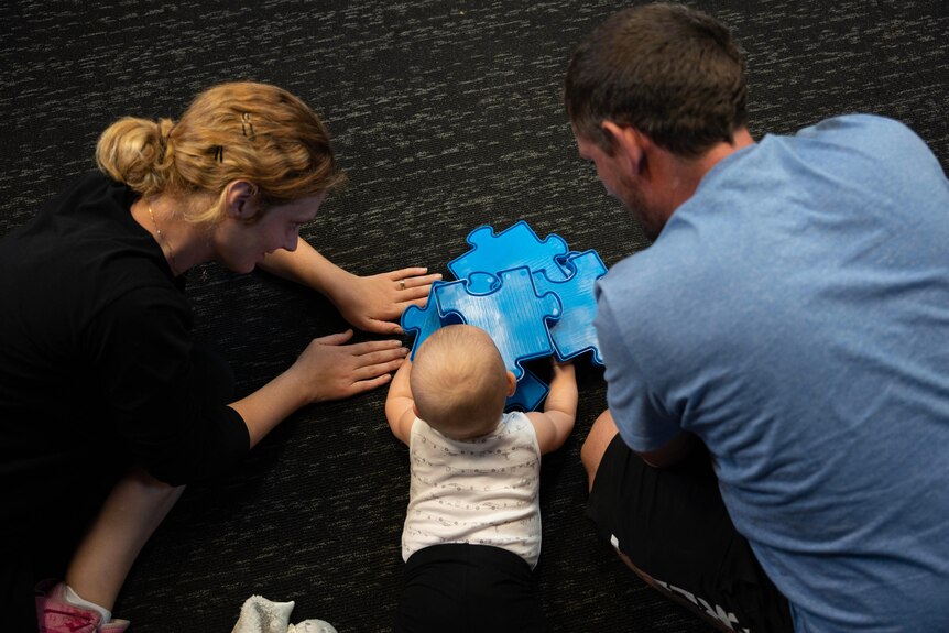 A man, woman and baby play with a jigsaw puzzle on the floor.