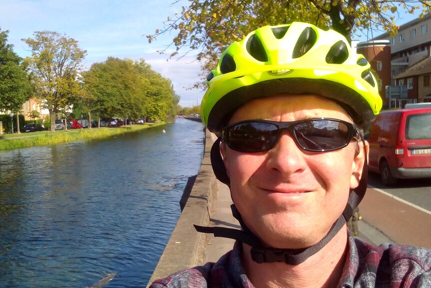 Chris Gray, wearing a bike helmet, smiles in a selfie taken by a canal on a sunny day.
