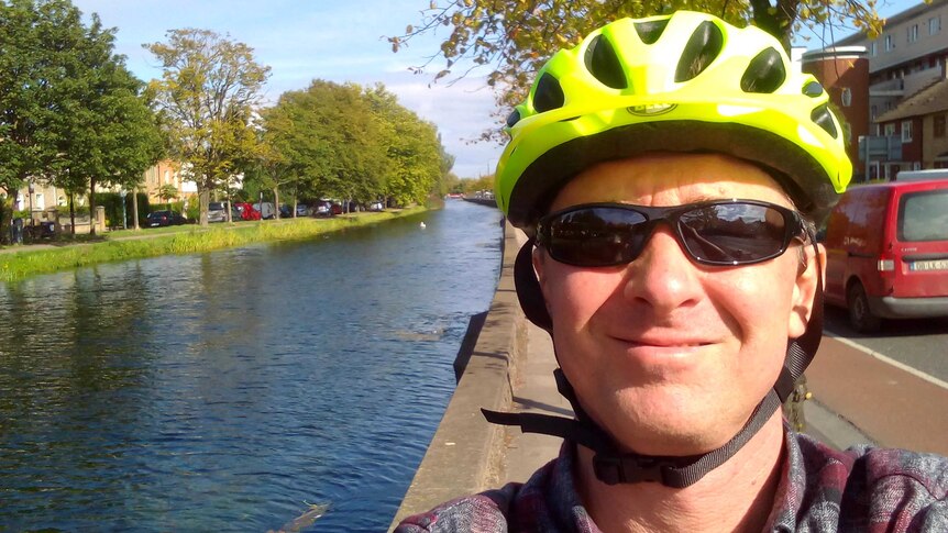 Chris Gray, wearing a bike helmet, smiles in a selfie taken by a canal on a sunny day.