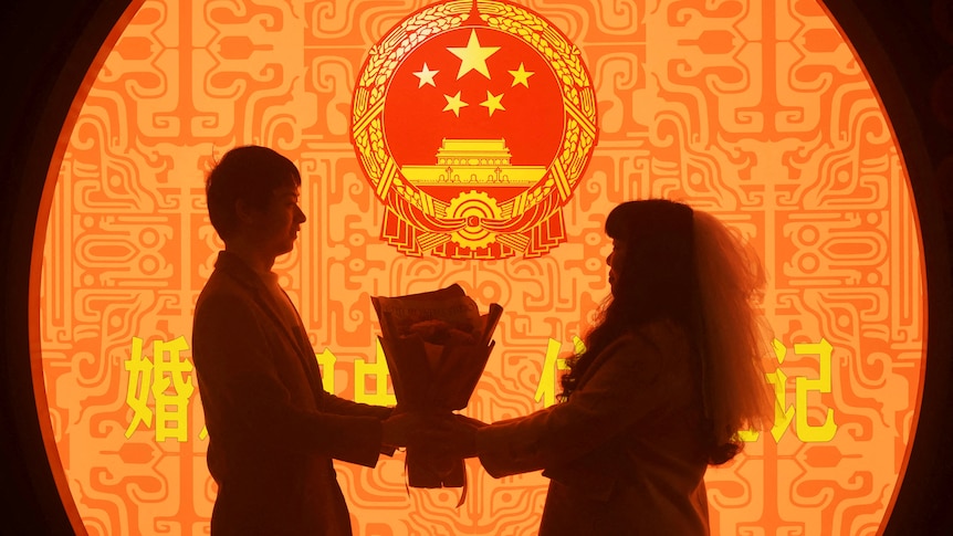 A couple pose in front of a brightly coloured wall with the national emblem of China, holding a bouqet of flowers between them