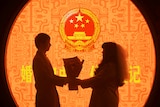 A couple pose in front of a brightly coloured wall with the national emblem of China, holding a bouqet of flowers between them