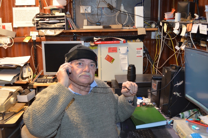 A man holds a phone to his ear while sitting at a desk in a cluttered workshop