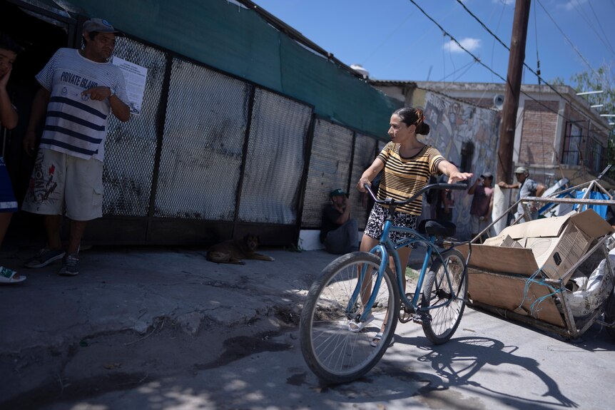 Una mujer joven se encuentra afuera de un edificio toscamente construido sosteniendo una bicicleta azul.