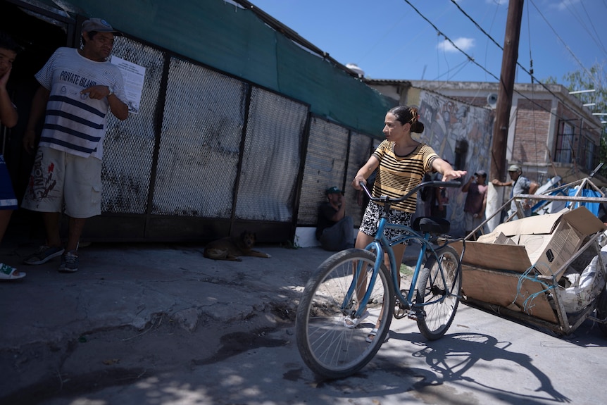 Молодая женщина стоит возле грубо построенного здания и держит в руках синий велосипед.