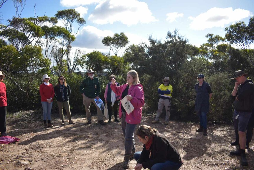 Mia Hunt wearing a pink jacket stands in a cleared area of bush surrounded by 9 onlookers.