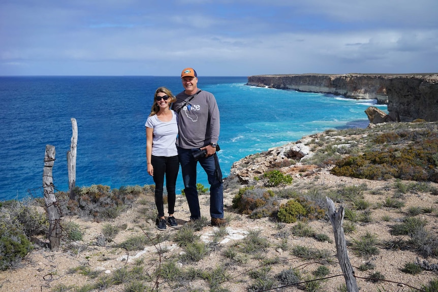 A woman and a man stand on a spectacular cliff, a deep blue ocean behind them, on a stunning day.