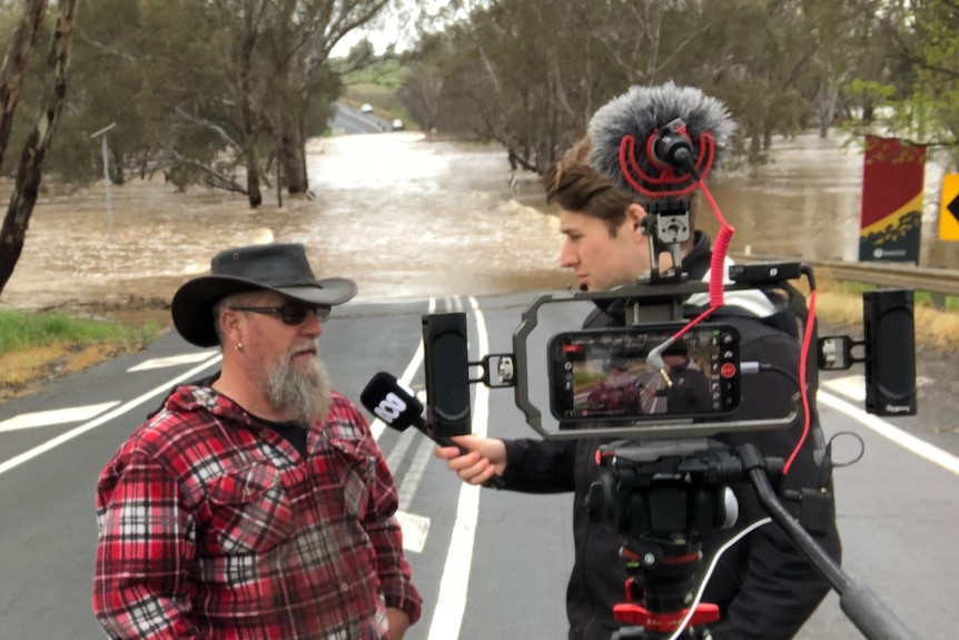 Man holding a microphone standing in front of a camera interviewing a man with flooded road in background.