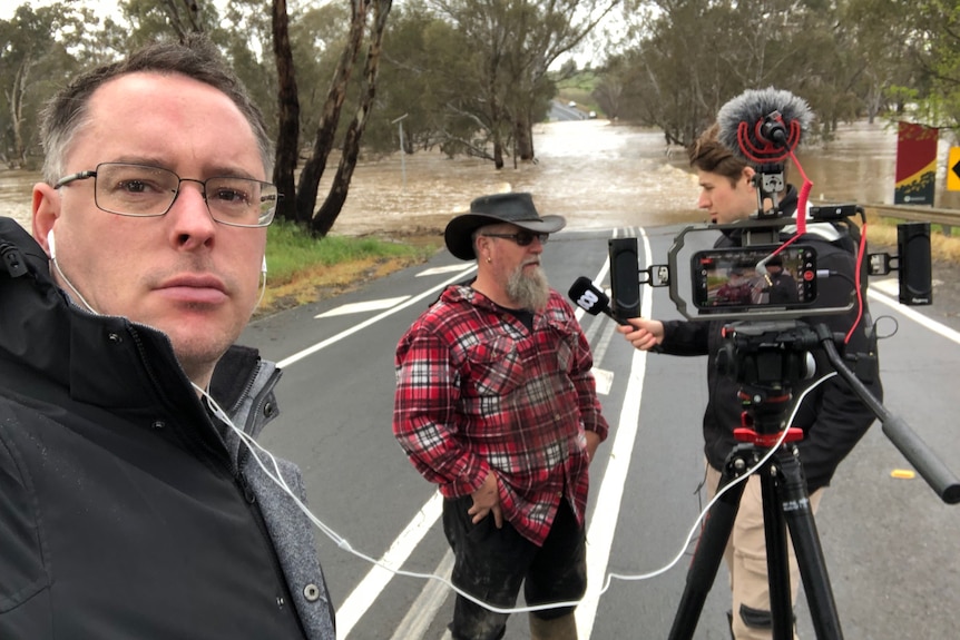 Man taking selfie of another man interviewing a farmer in front of a camera on a road cut by flood waters in background.