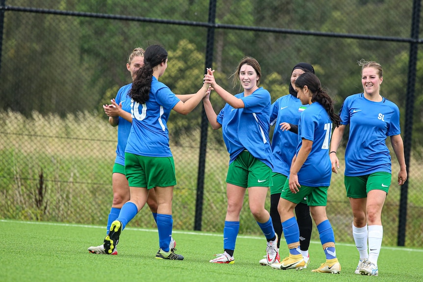A group of footballers celebrate after scoring a goal. Two women high five, while four other women come in to join in.