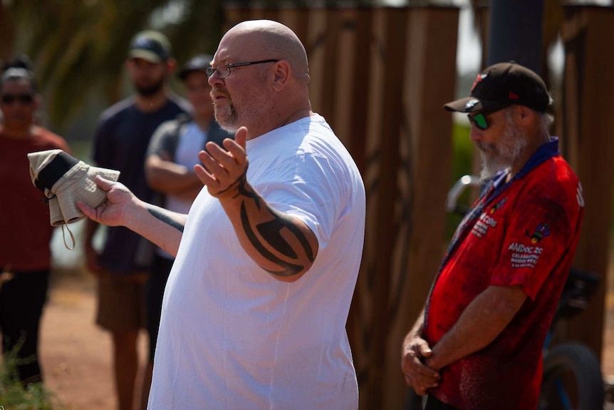 A bald man in a white shirt holds up his arms at an outdoor event.