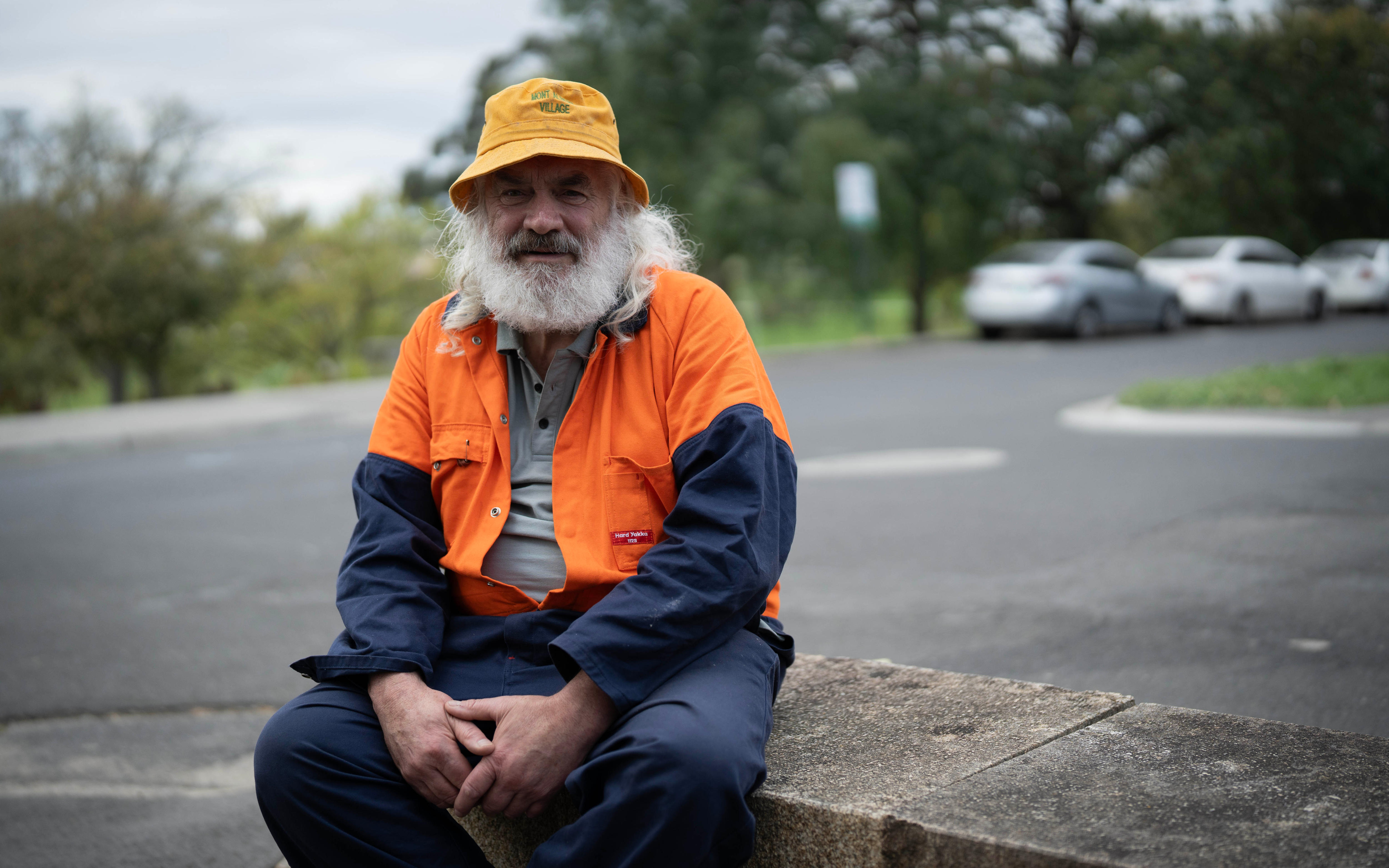 A man with a big white beard in a yellow bucket hat, orange and navy high vis jacket and pants sits on a street corner fence.