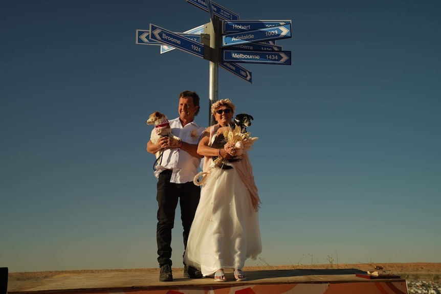 A bride and groom in front of a sign on a sand dune near Birdsville
