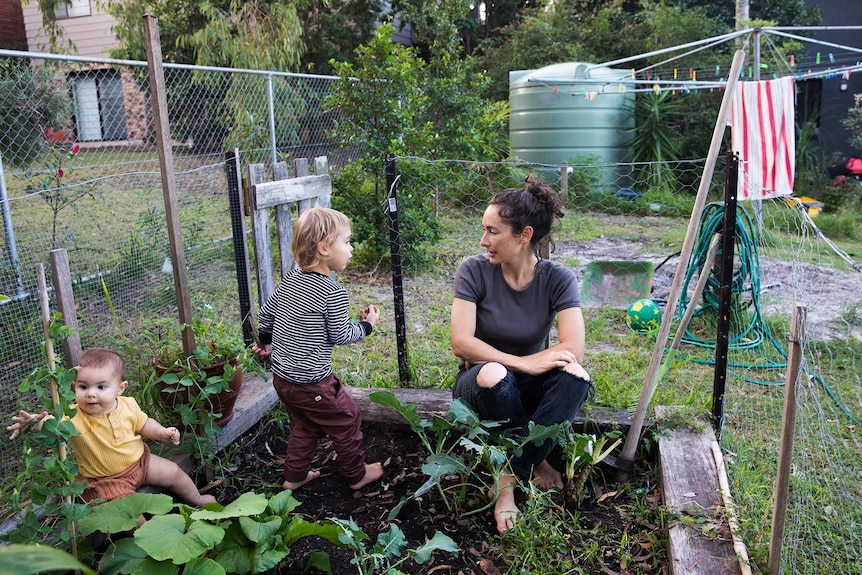 Milenka sitting in the veggie patch in their backyard with her two young kids.