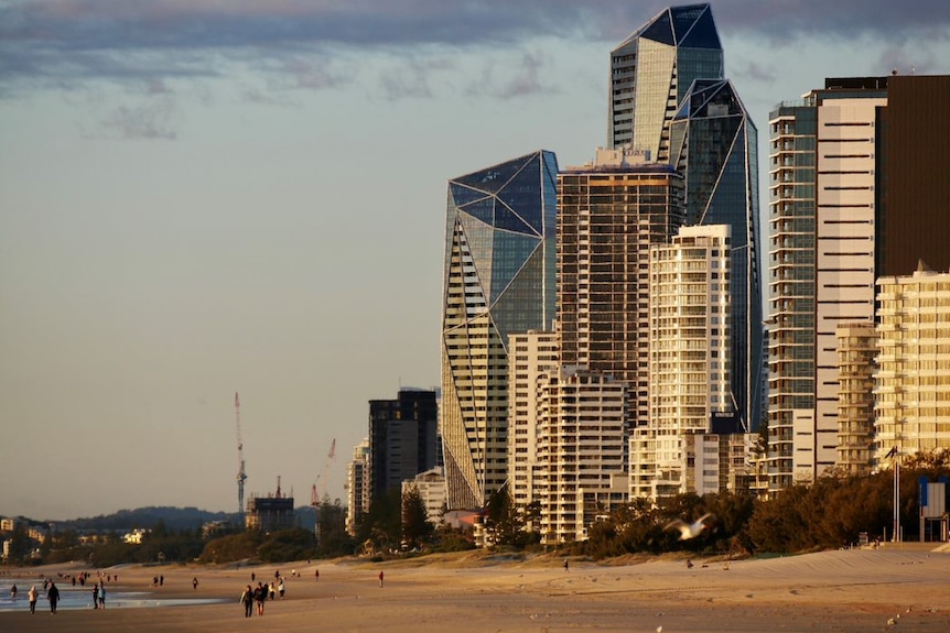 People in distance walk along a beach at dusk with skyscrapers and partly cloudy skies behind them.