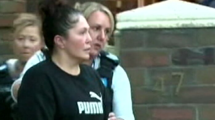 A woman wearing a black Puma jumper is escorted from her house by a police officer.
