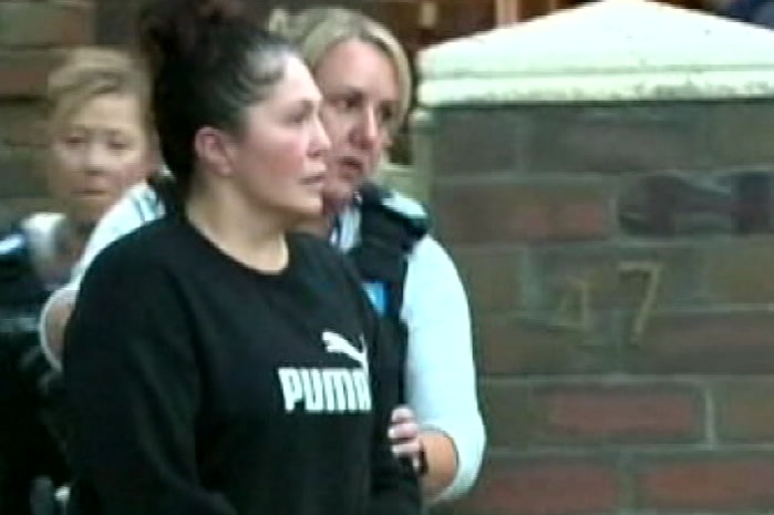 A woman wearing a black Puma jumper is escorted from her house by a police officer.