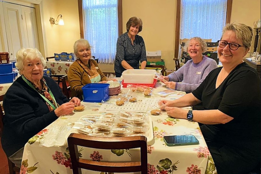 Six women sitting around a table and packaging biscuits look happily at the camera.