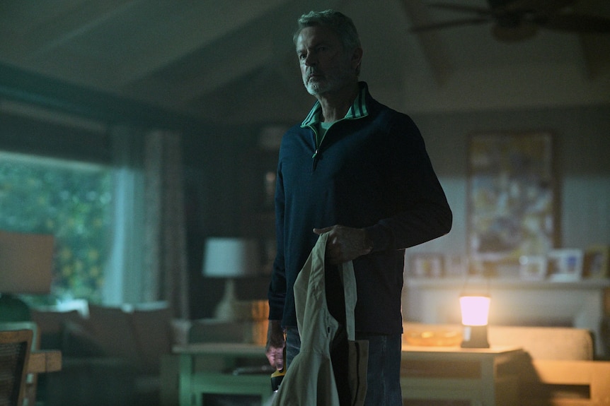 A TV still of Sam Neill standing in a shadowy living room, holding a jacket in one hand.