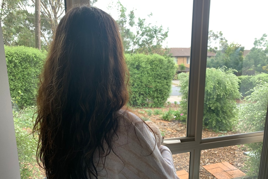 A woman with long dark hair shown from behind looks out a window.