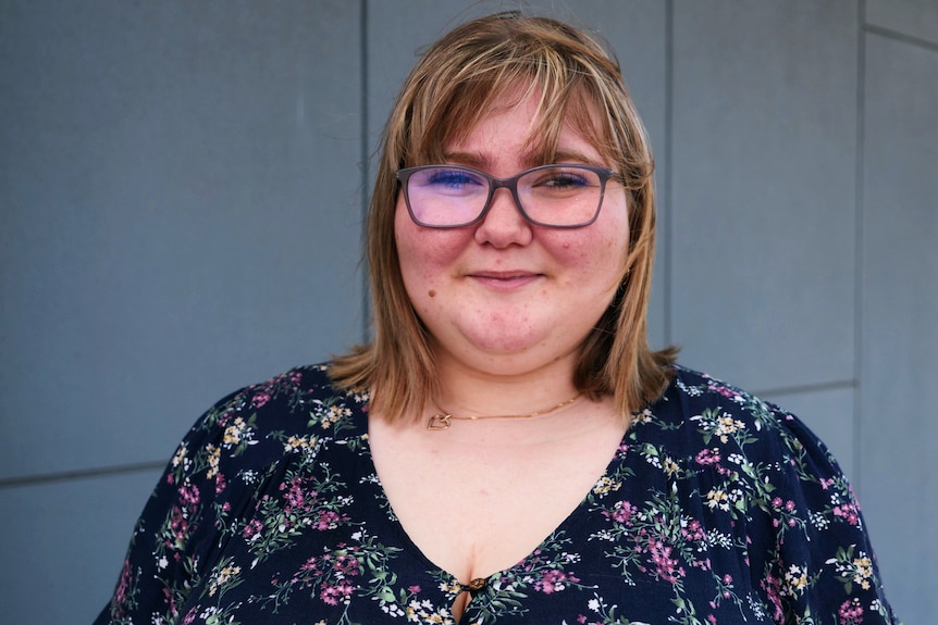 A woman in glasses smiles at the camera in front of a tiled wall.