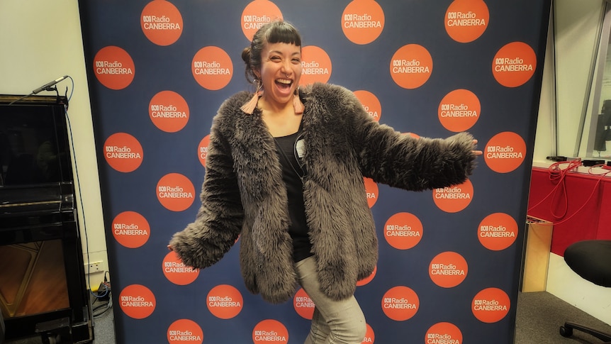 A happy woman in a radio studio, with arms outstretched and smiling, wearing a fur jacket and long earrings
