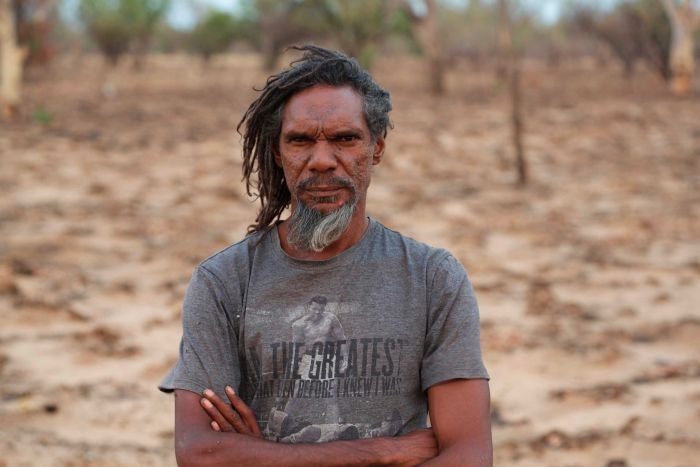 A serious-looking Indigenous Australian man stands with his arms folded looking at the camera