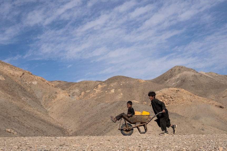 A boy pushes a wheelbarrow with canisters and his younger brother with hills and blue sky in the background