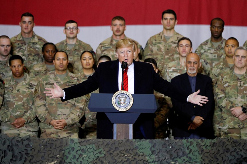 Donald Trump stands at a podium with his arms stretched out wide in front of a group of soldiers and next to a man.