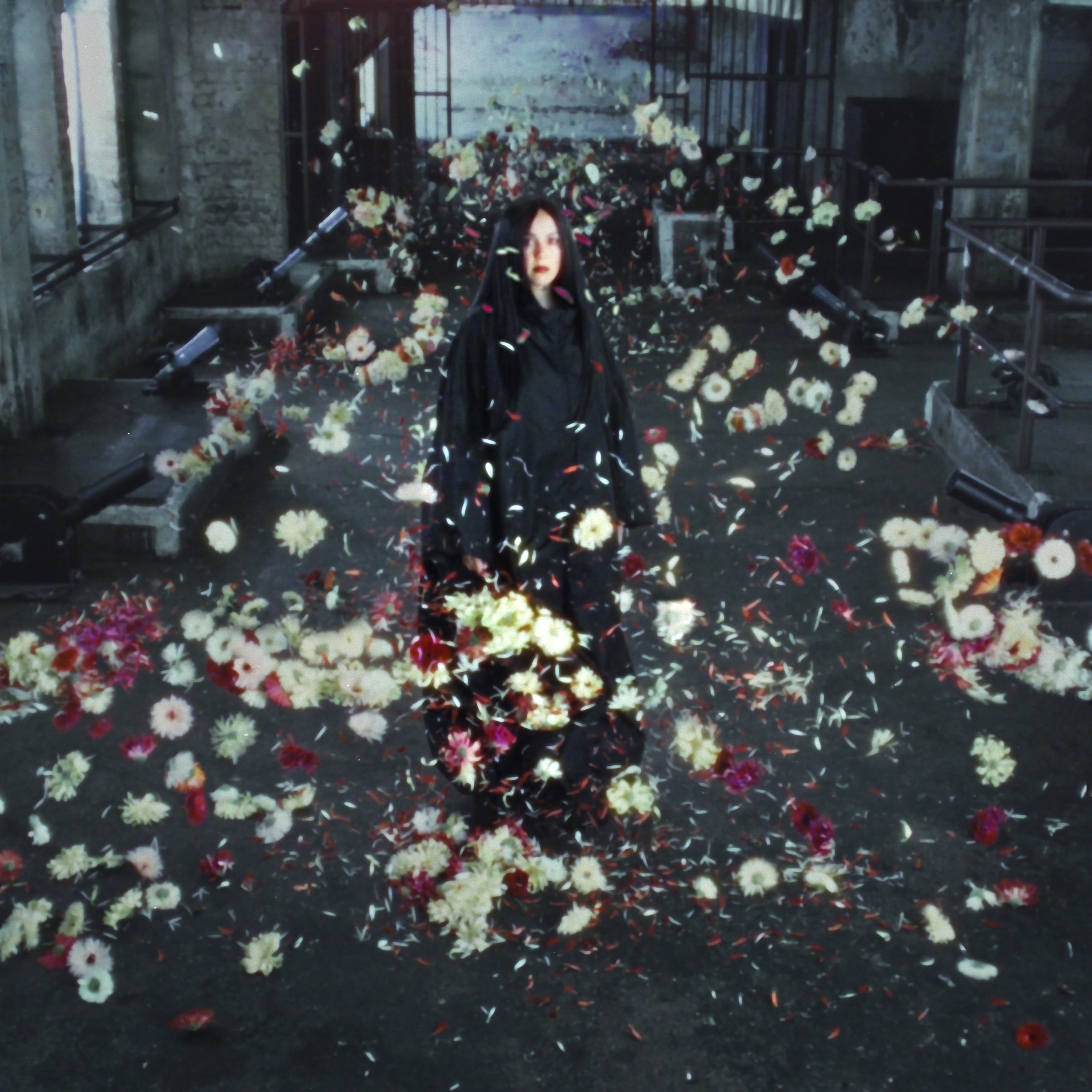 Woman stands in dark robe inside a stone mausoleum surrounded by an explosion of white daisies and petals