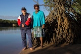 a man and a woman stand on the bank of a river with a mangrove to the left.