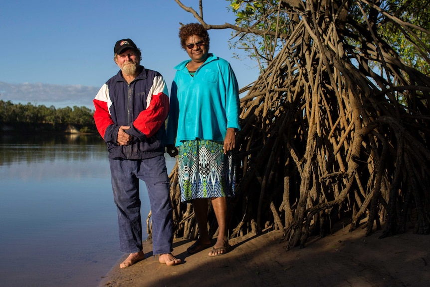 a man and a woman stand on the bank of a river with a mangrove to the left.