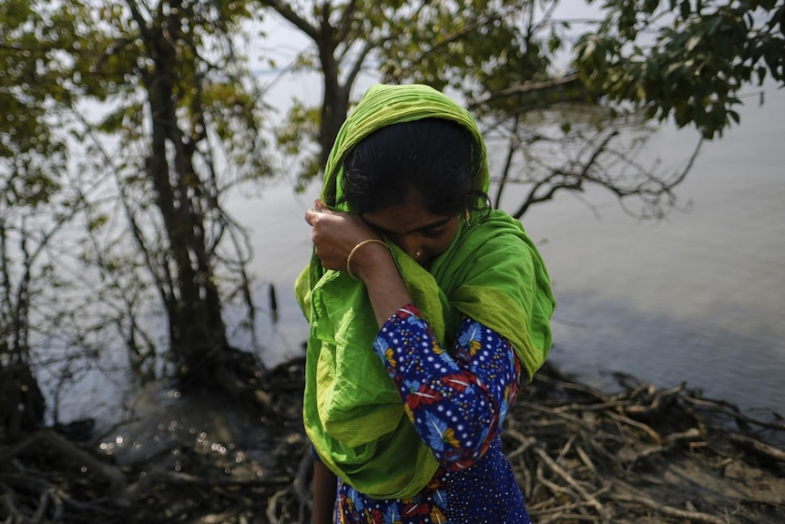 Reshma Begum, wearing a blue patterned dress and bright green shall wipes her tears as she stands on her lost land on mangrove 