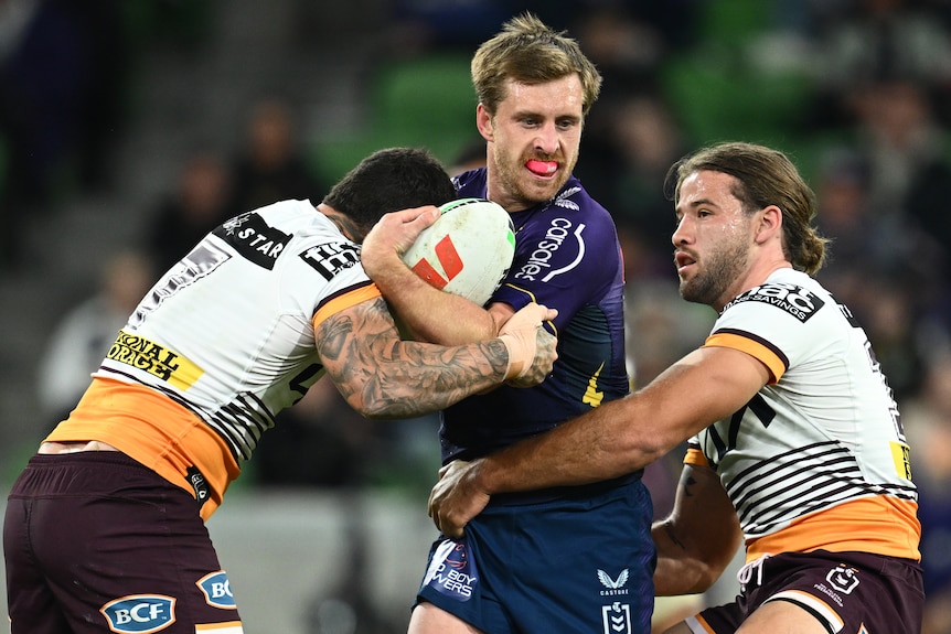 A Melbourne Storm NRL player holds the ball as he is tackled by two Brisbane Broncos opponents.
