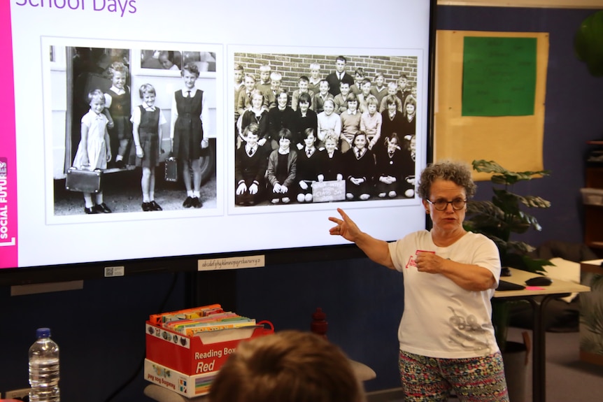 A woman with dwarfism wearing glasses standing in front of a projector screen with photos on it