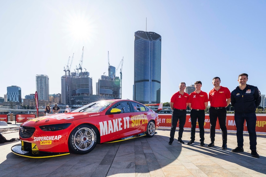 Four men stand on a rooftop with a red supercar.