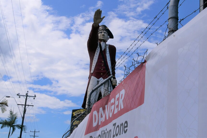 A large painted concrete statue of Captain Cook with his arm extended in front, stands behind a construction fence