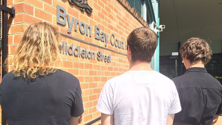 Three young men, facing a brick wall with the Byron Bay Court sign.