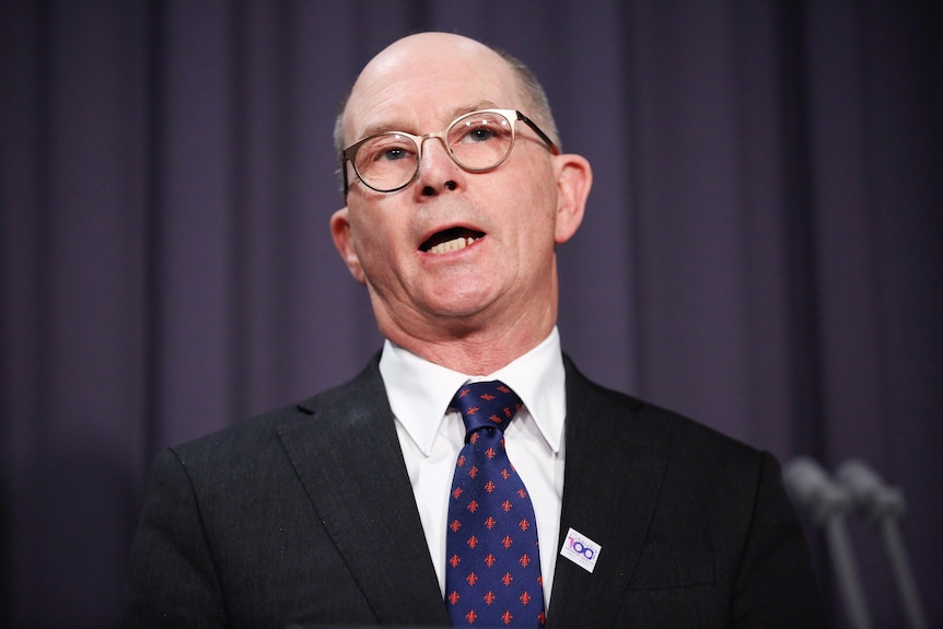 Kelly, a bald man with glasses, is mid-speech.