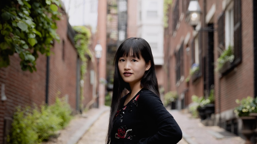 Rebecca F. Kuang standing outside in front of an apartment block, wearing black, looking at the camera